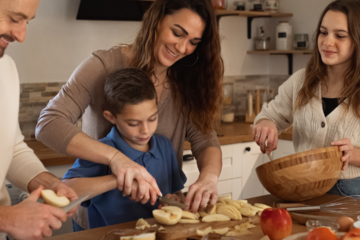 Famille qui cuisine ensemble dans une cuisine chaleureuse : une femme aide son fils à couper des pommes, le père et la fille préparent le repas, illustrant le rééquilibrage alimentaire au quotidien – cabinet Luxo'Santé à Challans