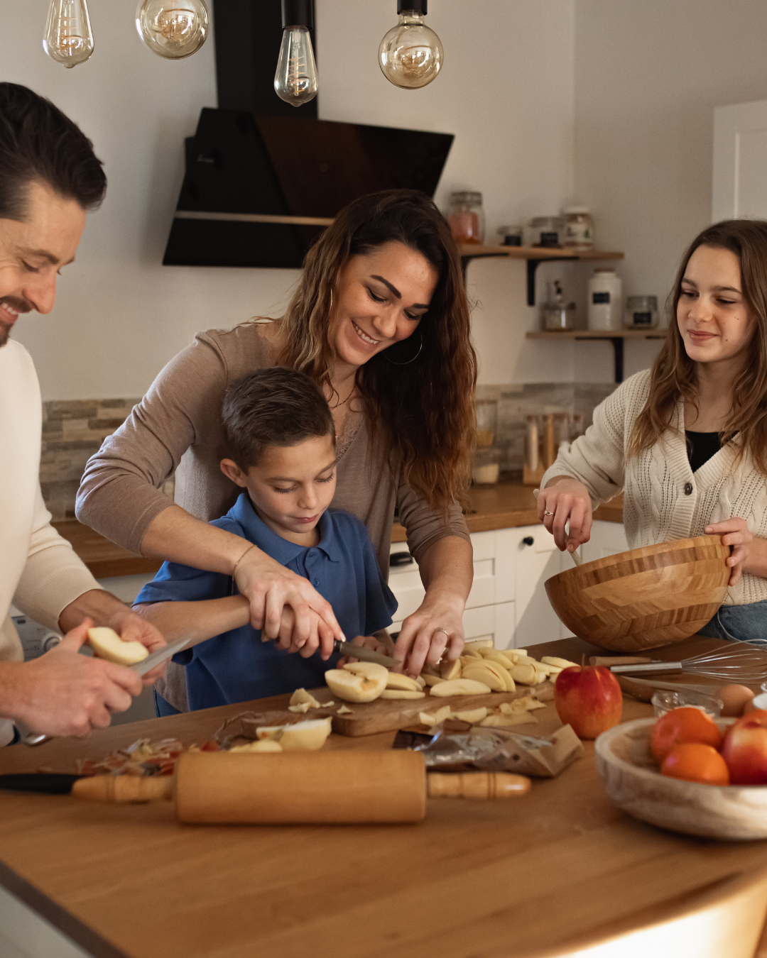 Famille qui cuisine ensemble dans une cuisine chaleureuse : une femme aide son fils à couper des pommes, le père et la fille préparent le repas, illustrant le rééquilibrage alimentaire au quotidien – cabinet Luxo'Santé à Challans